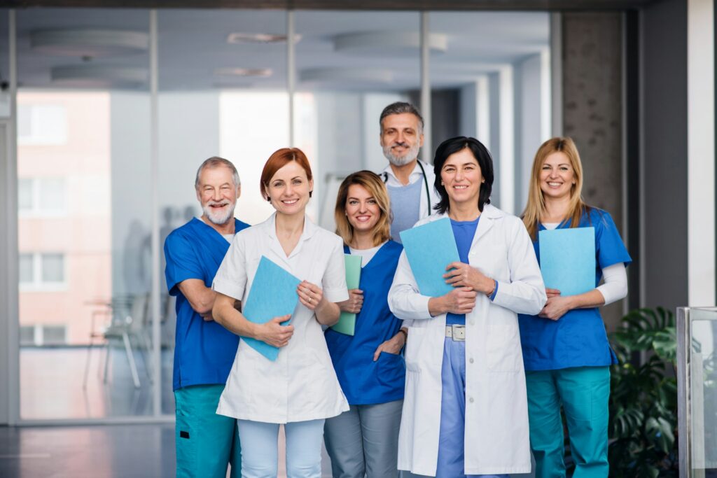 Six smiling clinicians in scrubs and white coats stand in a glass-walled corridor, most holding blue or green folders