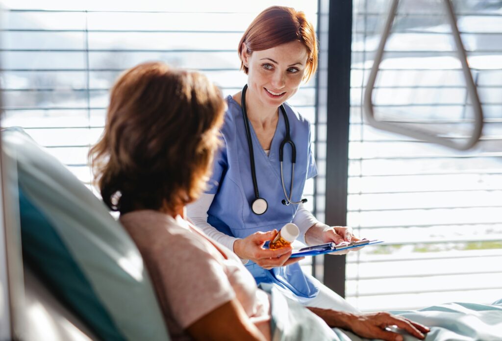 Pill bottle and clipboard at a bedside | Clinical Match Me Smiling clinician in blue scrubs holds a pill bottle and a clipboard beside a woman sitting up in a hospital bed