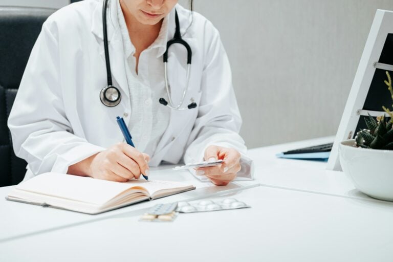 Doctor in a white coat with a stethoscope around the neck writes in a notebook at a white desk, with a calculator and papers nearby.