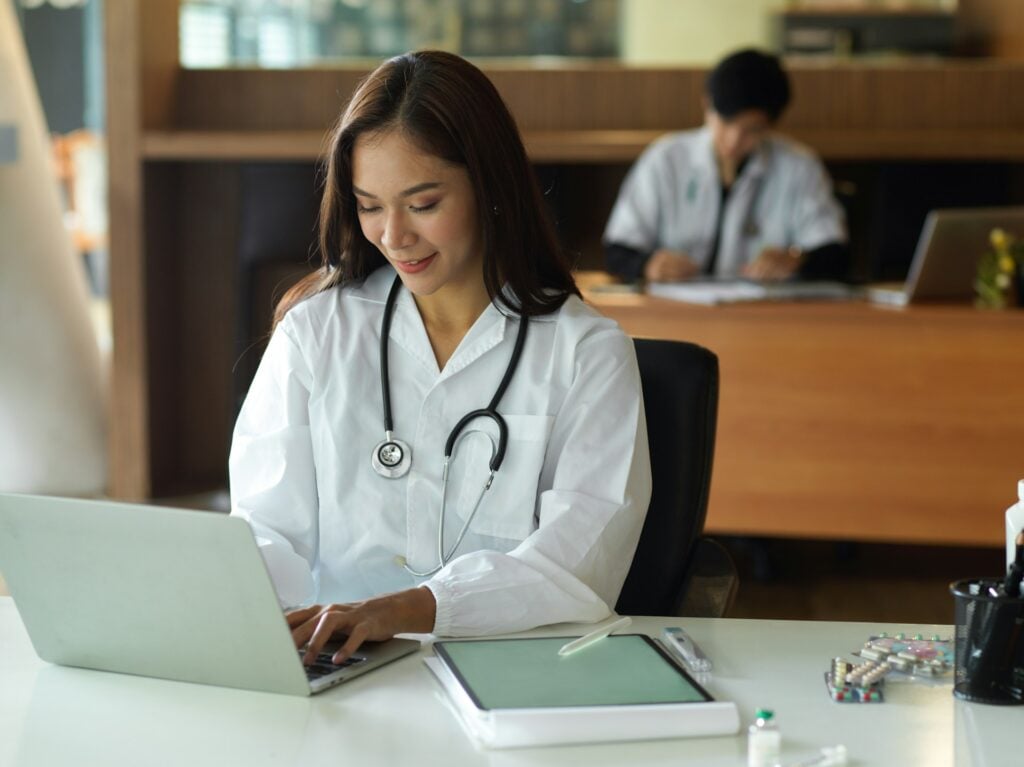 Clinician typing in a shared office | Clinical Match Me At her desk, a woman in a white coat and stethoscope smiles as she types on a laptop, with a colleague at work behind her
