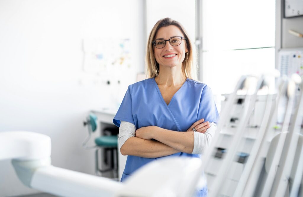 Smiling woman in a treatment room | Clinical Match Me Woman in glasses and blue scrubs smiles with arms folded in a bright treatment room