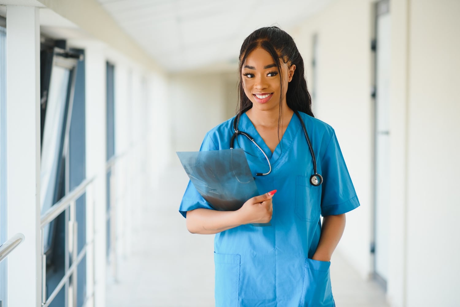 Female healthcare professional in blue scrubs holding an X-ray film in a bright hospital hallway, stethoscope around her neck.