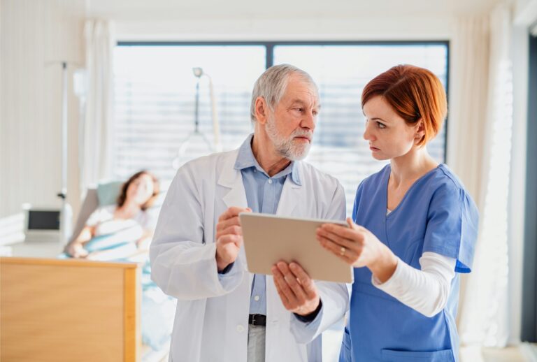 Clinicians conferring beside a hospital bed | Clinical Match Me Gray-haired clinician in a white coat and a colleague in blue scrubs confer over a tablet near a patient resting in bed
