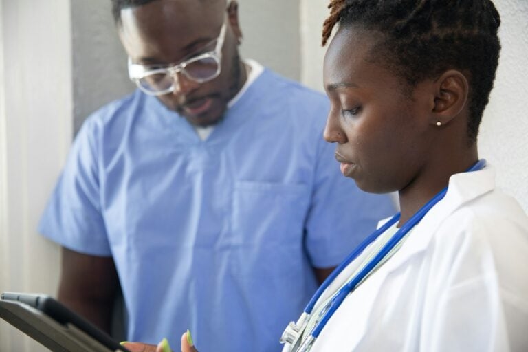Colleagues looking at a tablet | Clinical Match Me While a colleague in light blue scrubs and clear glasses looks on, a clinician in a white coat reads a tablet