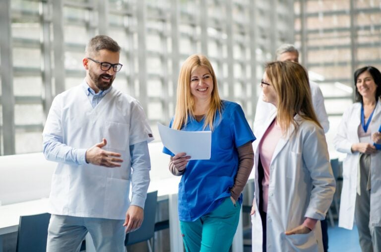Group chatting in a sunlit corridor | Clinical Match Me Bearded man in glasses gestures as he talks with smiling colleagues in white coats and scrubs in a sunlit corridor
