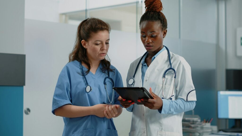 Stethoscopes around their necks, two clinicians in blue scrubs and a white coat study a tablet together