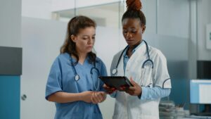 Two healthcare professionals review data on a tablet in a clinical setting, one in blue scrubs and the other in a white coat with stethoscopes nearby.