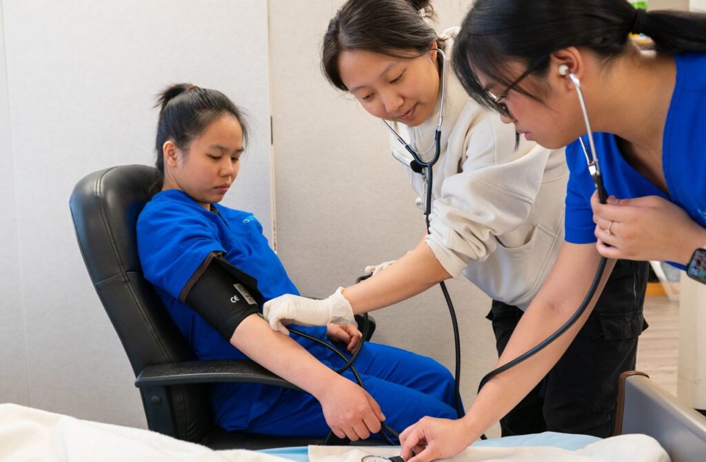 Taking blood pressure in a clinic room | Clinical Match Me Clinician in a cream sweater takes the blood pressure of a seated woman in blue scrubs as a colleague listens