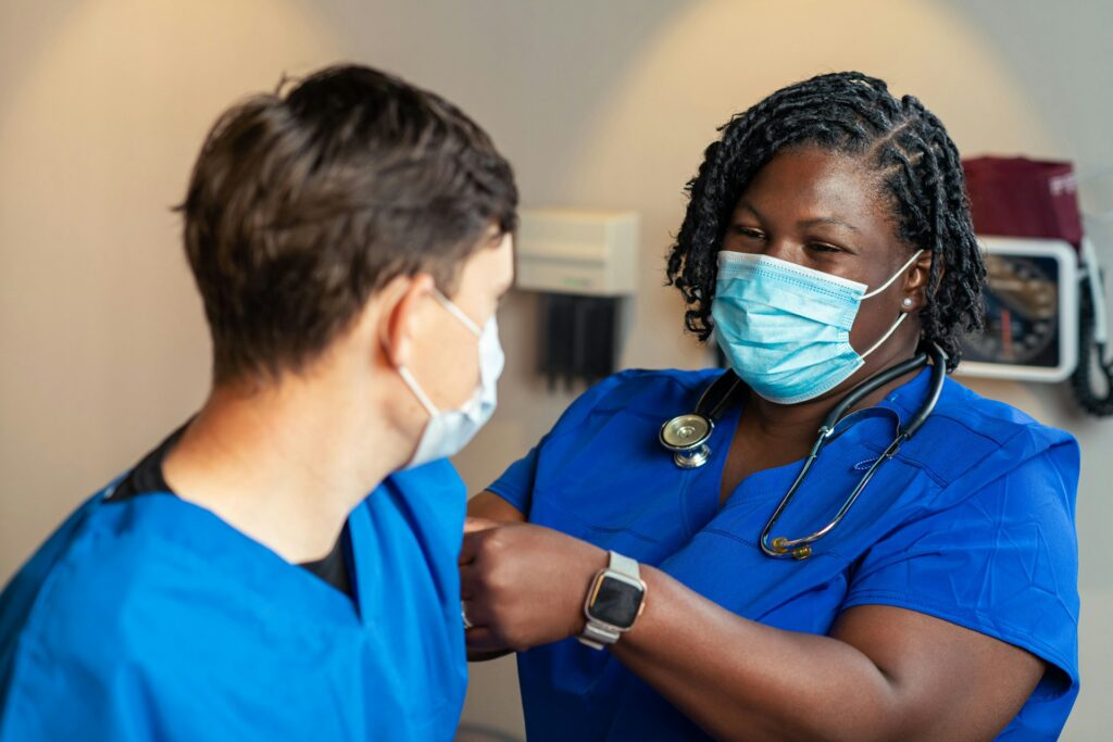 Masked clinician in an exam room | Clinical Match Me Masked clinician with braids and a stethoscope places a hand on the arm of a masked person in blue scrubs in an exam room