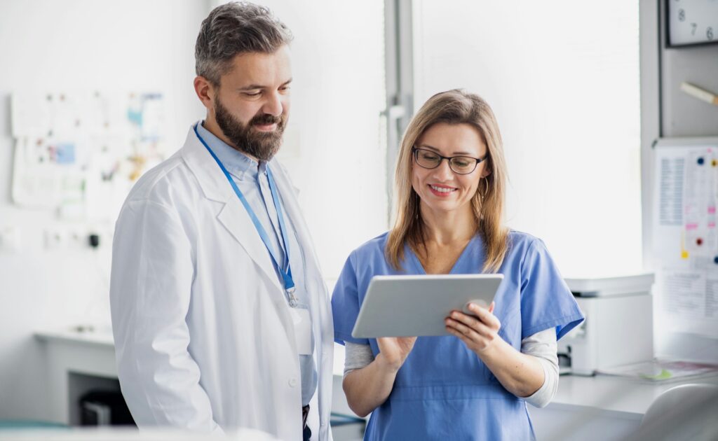 Clinician showing a colleague a tablet | Clinical Match Me Clinician in blue scrubs shows a tablet to a bearded colleague in a white coat in a bright clinic office