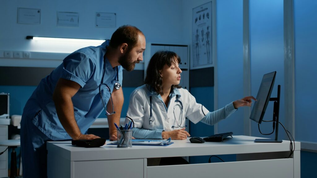 Colleagues reviewing a computer screen | Clinical Match Me In a dimly lit office, a colleague in a white coat points at a monitor as a bearded clinician in blue scrubs leans in