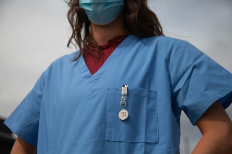 Masked person in blue scrubs | Clinical Match Me Low-angle view of a person in a blue surgical mask and blue scrubs, hands on hips, a badge reel clipped to the pocket
